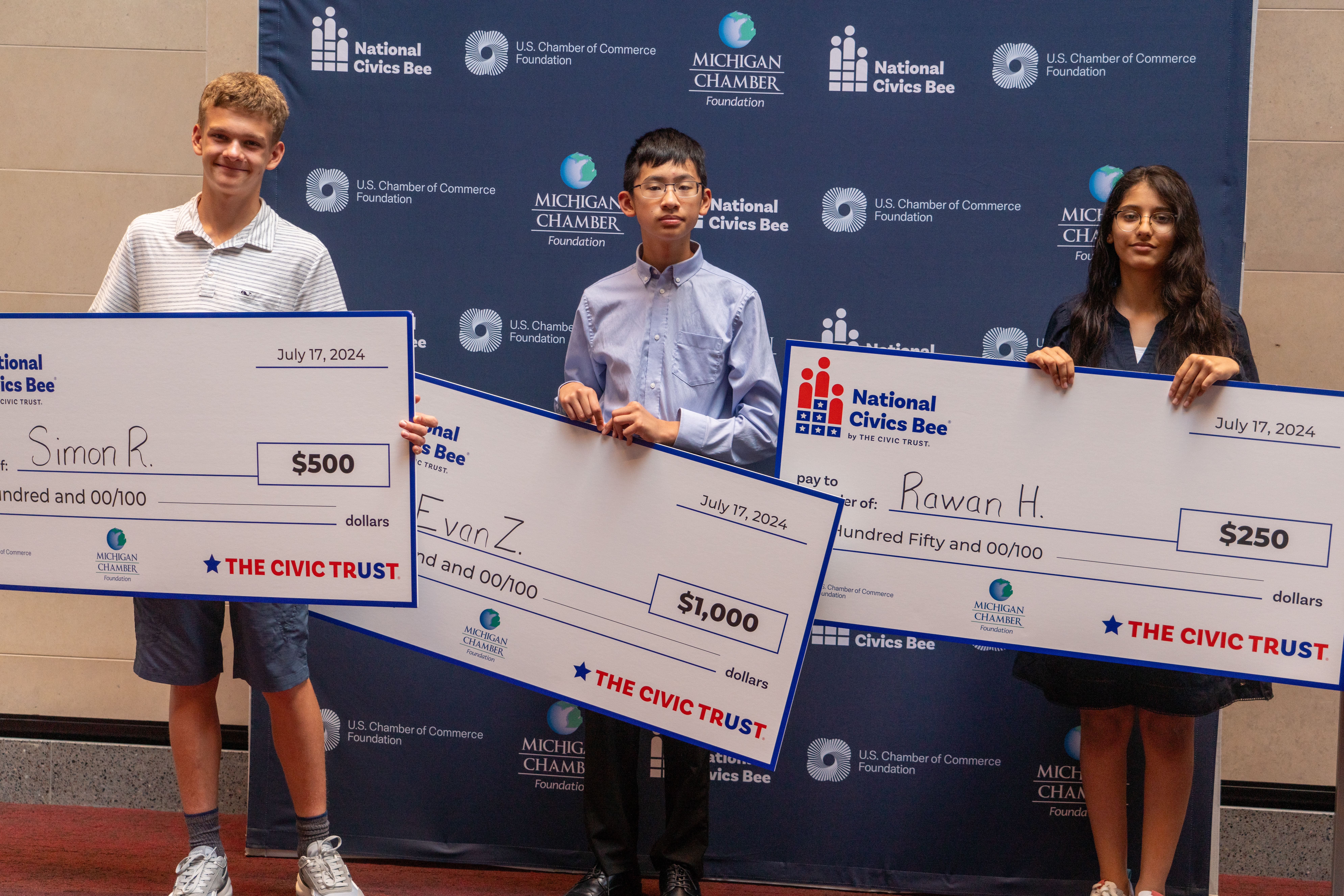 Three students proudly hold oversized checks as they stand in front of a backdrop featuring logos of the National Civics Bee, U.S. Chamber of Commerce Foundation, and Michigan Chamber Foundation. The student on the left, Simon R., is smiling and holding a check for $500. In the center, Evan Z. stands with a serious expression, holding a check for $1,000. On the right, Rawan H. holds a check for $250. All three students are dressed in casual attire, and the backdrop reinforces the official nature of the event. The date on the checks reads July 17, 2024.