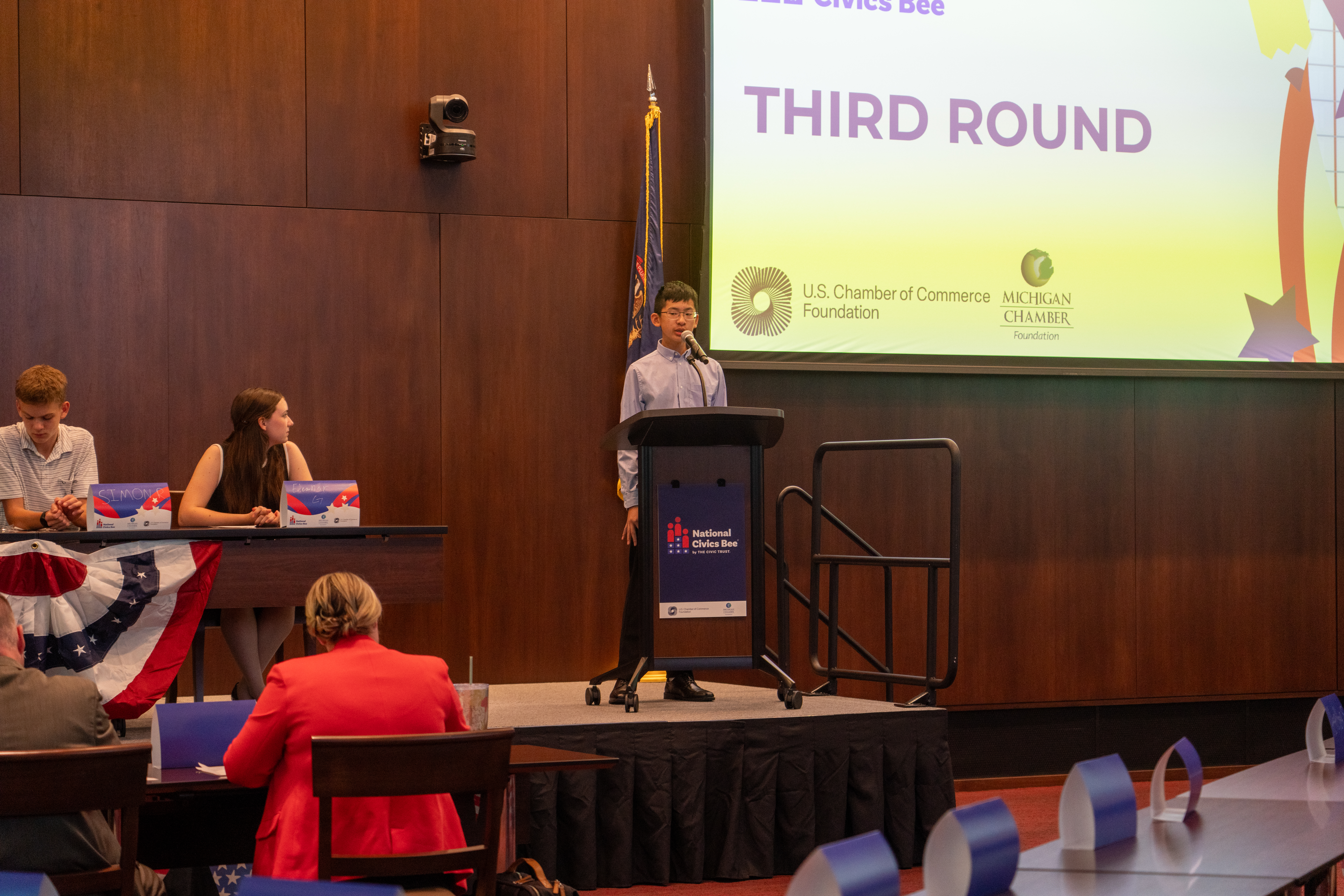 A young participant stands confidently behind a podium during the third round of the National Civics Bee competition. The event is hosted in a formal setting, with a large screen in the background displaying the words 