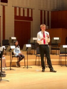 A young student stands on stage holding a microphone, presenting confidently during a competition. He is dressed in a white shirt, black pants, and a red tie, giving a formal and polished appearance. Behind him, another participant sits on a chair, listening attentively. The stage is set in a large hall with a wooden backdrop and music stands placed around, indicating an academic or formal setting. The setting suggests a public speaking or debate event, with other participants visible in the background.