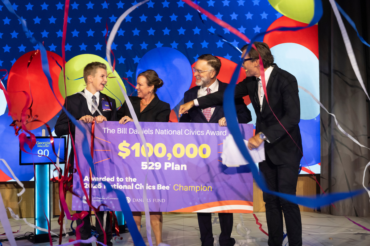 A group of four people on stage celebrate as they unveil a large ceremonial check for $100,000 awarded to the 2026 National Civics Bee Champion.
